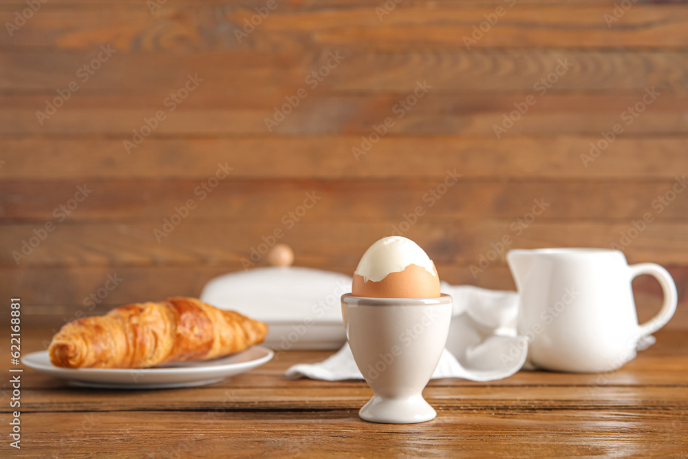 Holder with boiled chicken egg and plate of croissant on wooden background