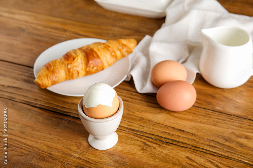 Holder with boiled chicken egg and plate of croissant on wooden background