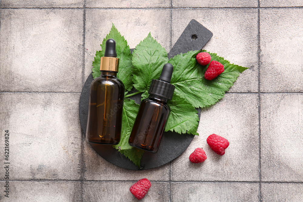 Board with bottles of cosmetic raspberry oil on grey tile background