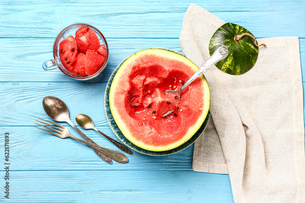 Half of fresh watermelon and cup with pieces on blue wooden background