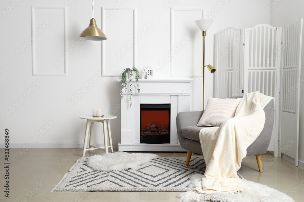 Cozy grey armchair with cushion, fireplace and folding screen in interior of living room