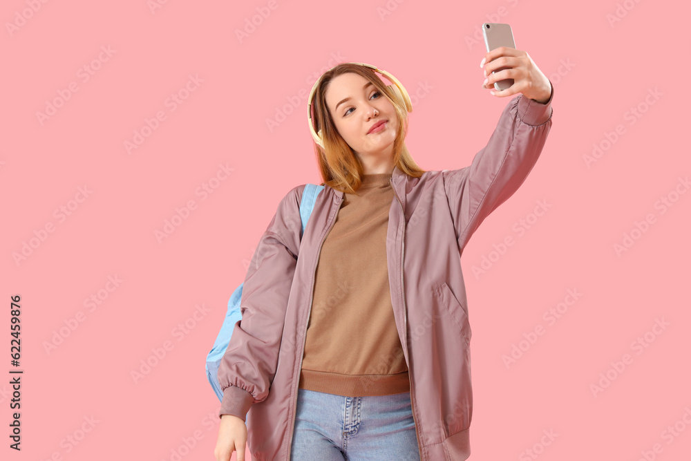 Female student in headphones with mobile phone taking selfie on pink background