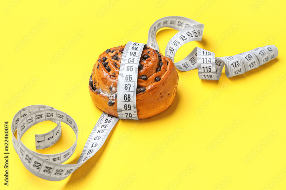 Tasty chocolate bun and measuring tape on yellow background, closeup. Diet concept