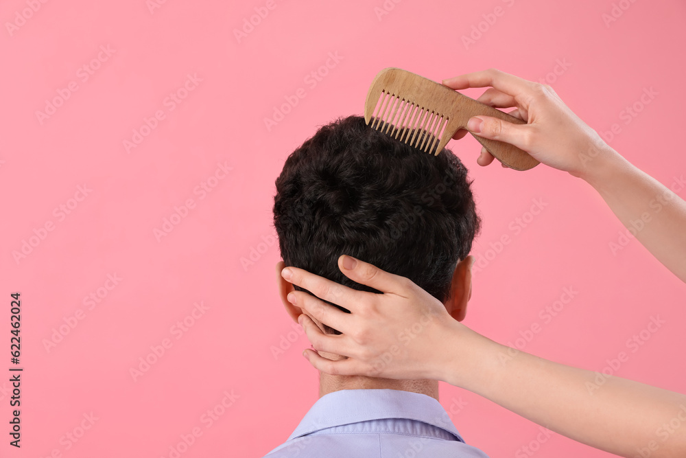 Female hairdresser combing hair of young man on pink background, back view