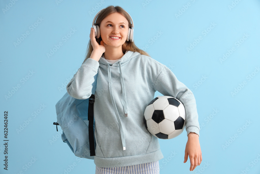 Female student with backpack and soccer ball listening music in headphones on blue background