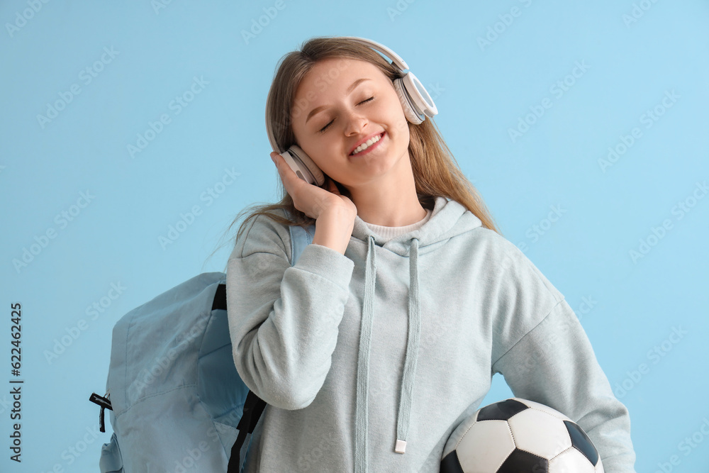 Female student with backpack and soccer ball listening music in headphones on blue background