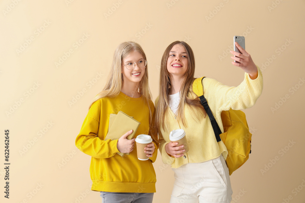 Happy female students with books and cups of coffee taking selfie on pale yellow background