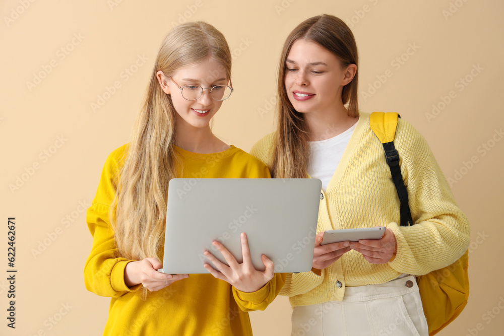 Happy female students with laptop and tablet on pale yellow background