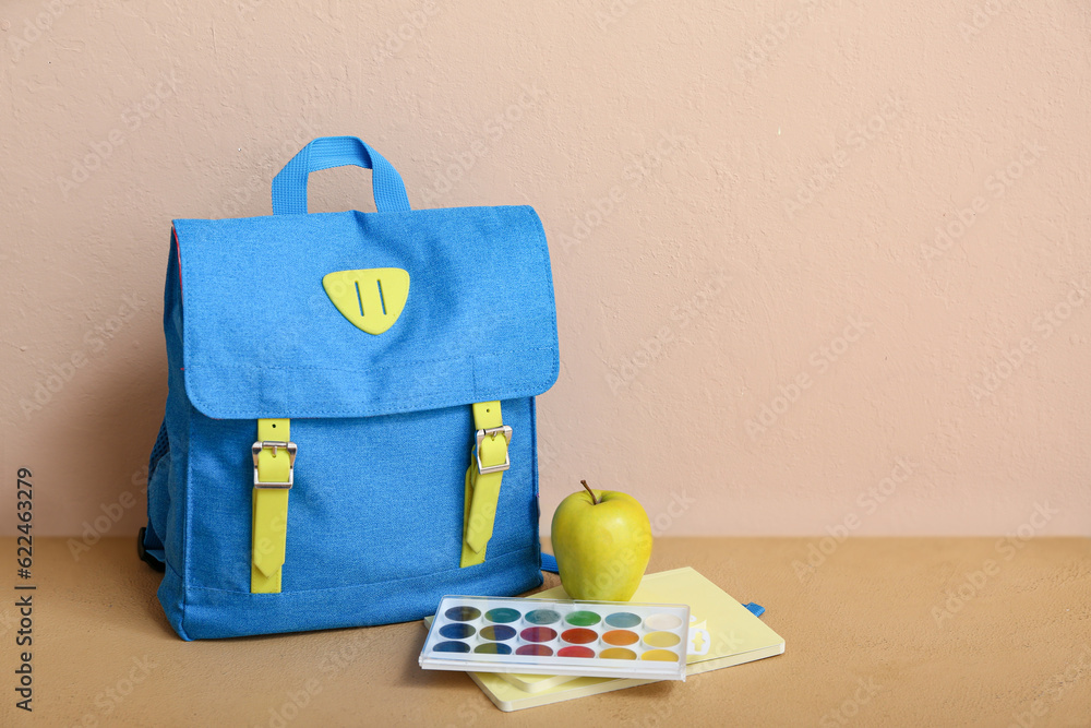 Blue school backpack with apple, watercolor and notebook on table near beige wall