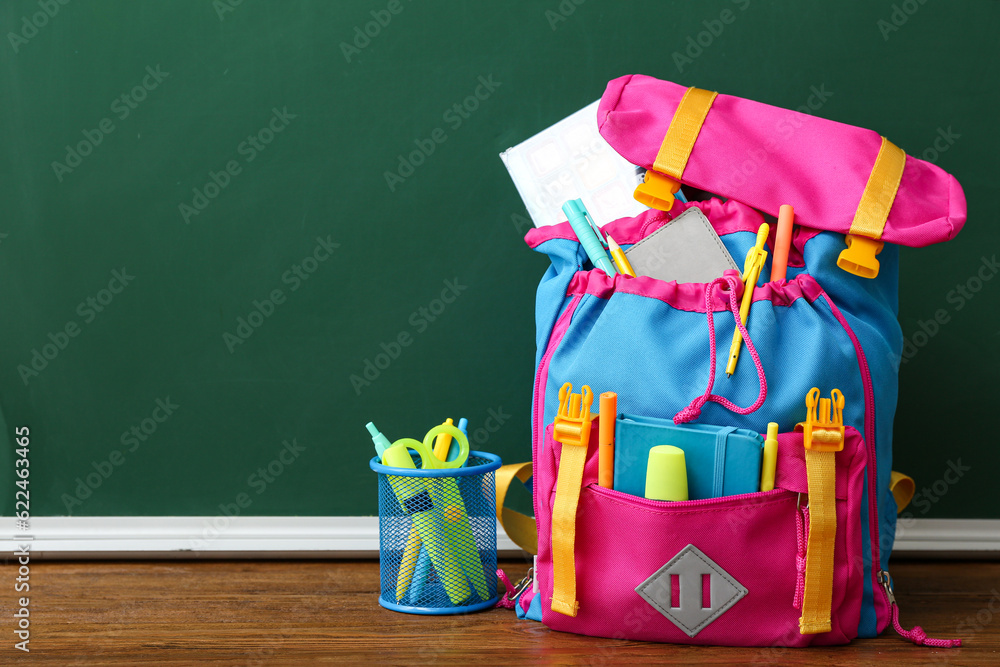 Color school backpack with markers, pens and watercolor on brown wooden table near chalkboard