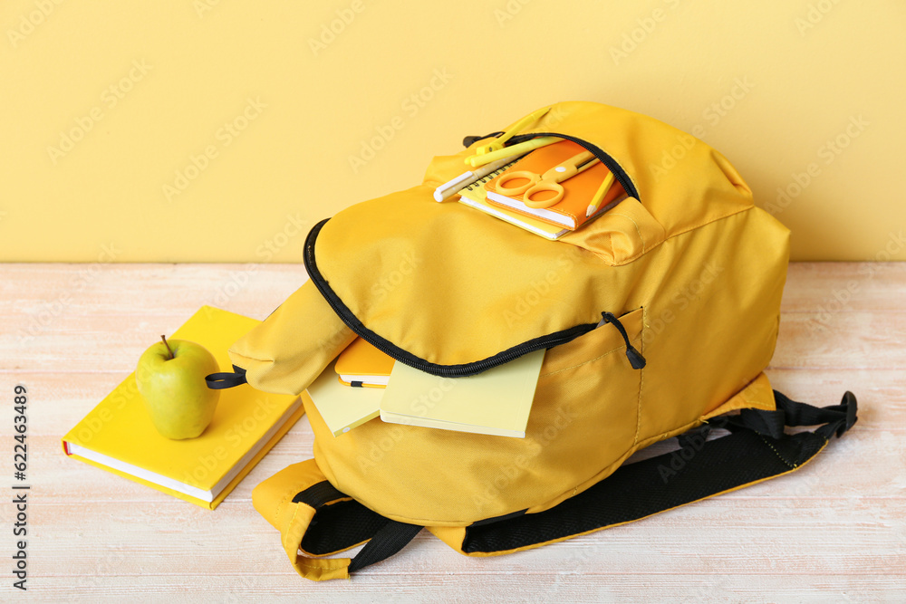 Yellow school backpack with notebooks, apple and scissors on wooden table near wall