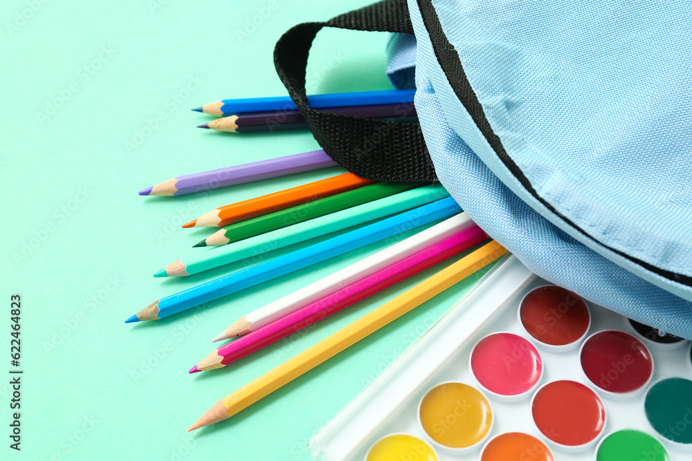 School backpack with different colorful pencils and paints on turquoise background, closeup