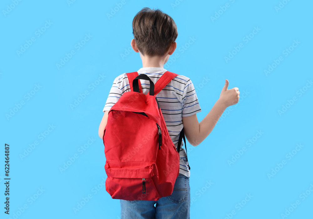 Little boy with schoolbag showing thumb-up on blue background, back view