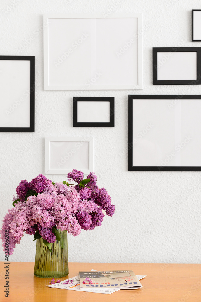 Vase with beautiful lilac flowers, magazines on table and blank pictures, closeup