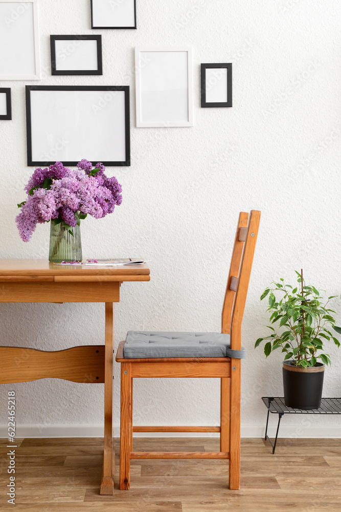 Vase with beautiful lilac flowers on table, chair and blank pictures in interior of light living roo