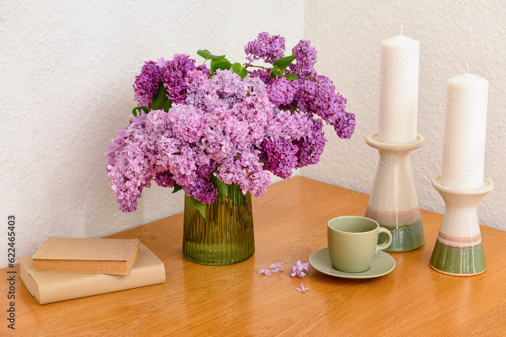 Vase with beautiful lilac flowers, candles, cup and books on table near light wall in room, closeup