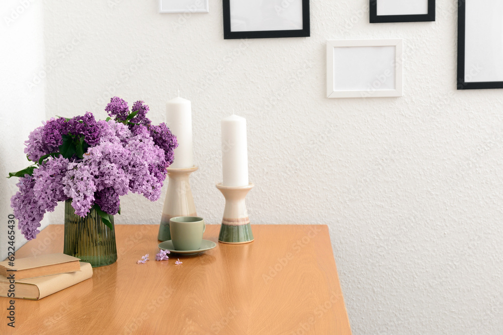 Vase with beautiful lilac flowers, candles on table and blank pictures in interior of light living r