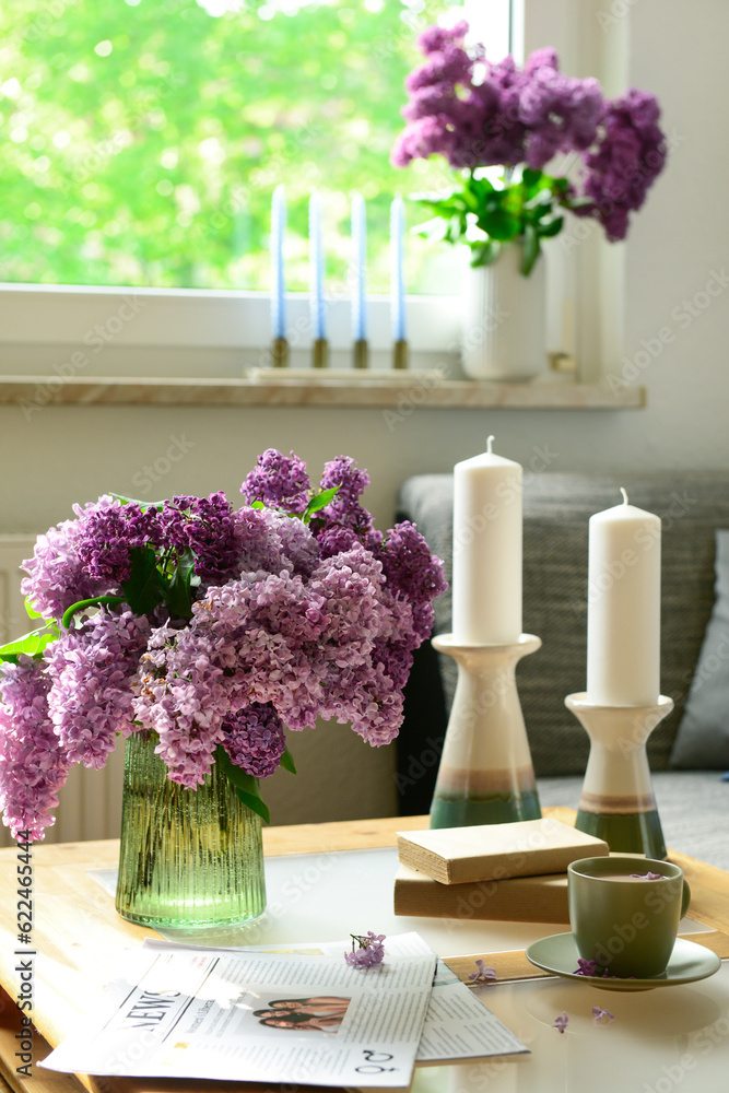 Vase with beautiful lilac flowers, candles, cup and newspaper on table in interior of light living r