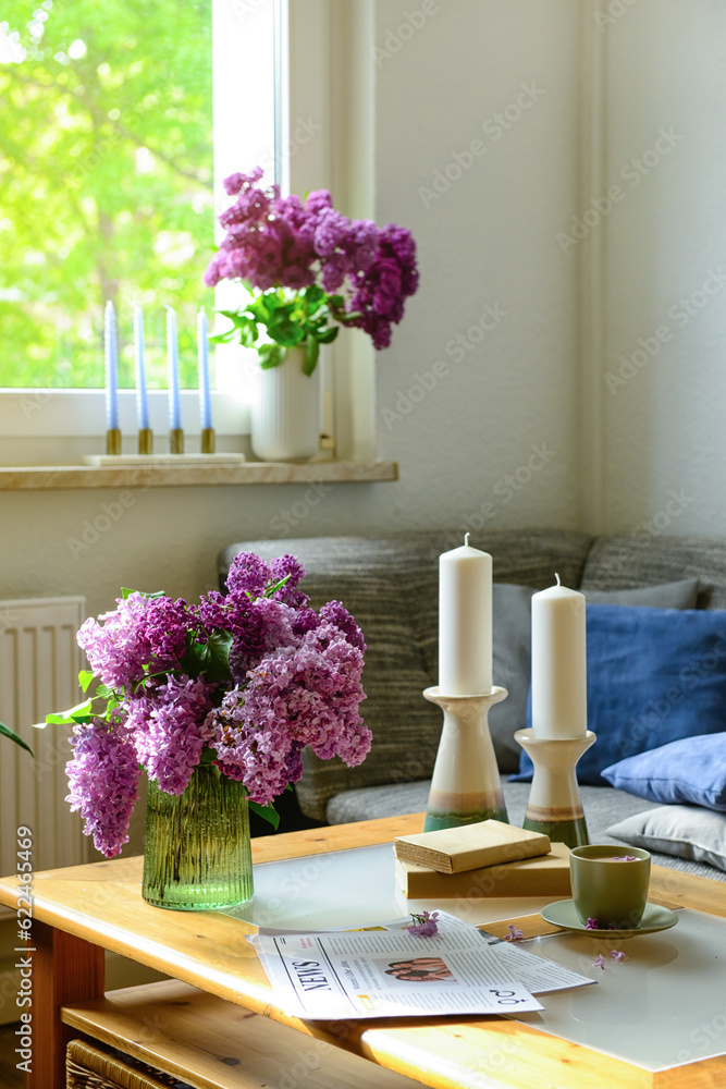 Vase with beautiful lilac flowers, candles, cup and newspaper on table in interior of light living r