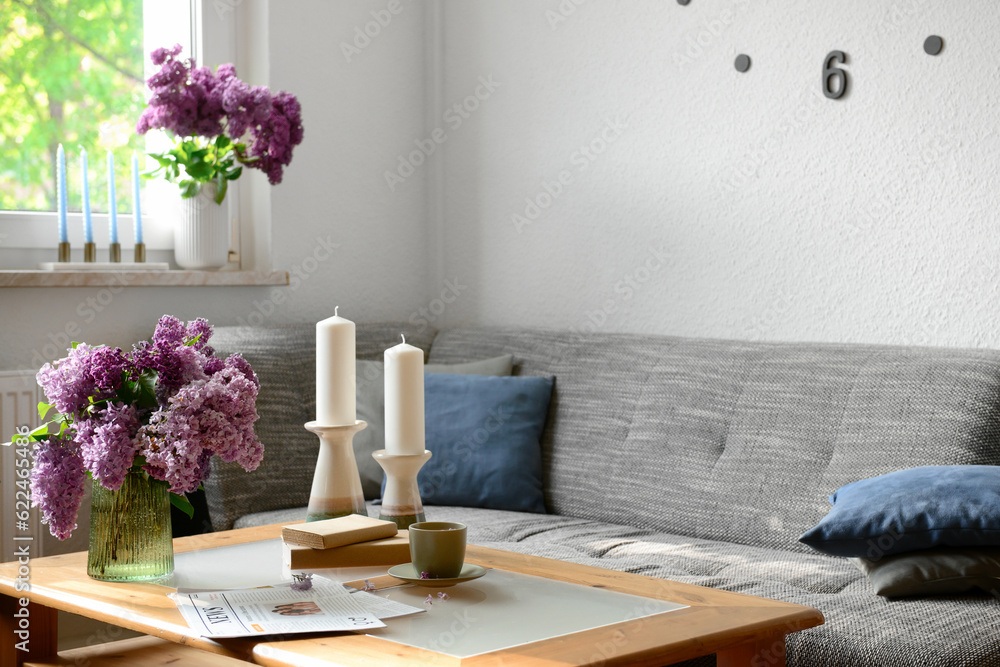 Interior of light living room with beautiful lilac flowers, candles, table and sofa