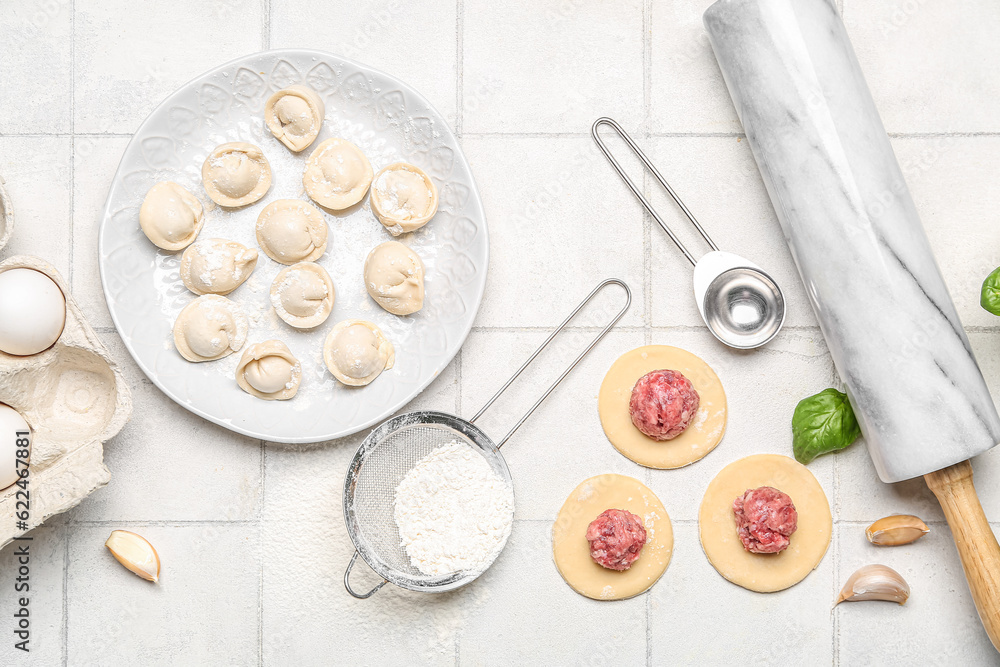 Plate with uncooked dumplings and ingredients on white tile background