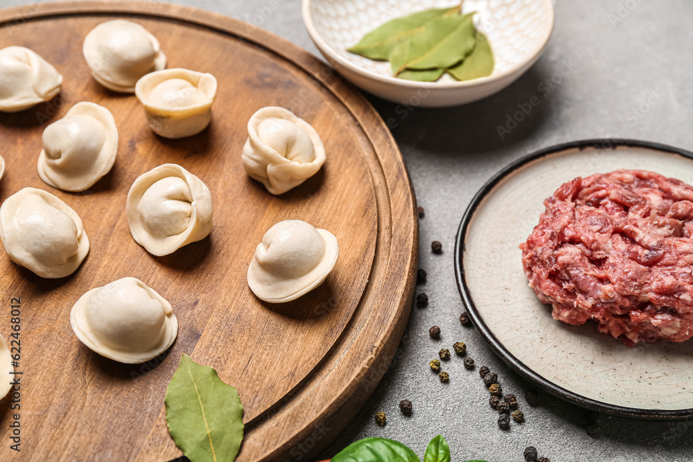 Wooden board with uncooked dumplings and minced meat on grey background