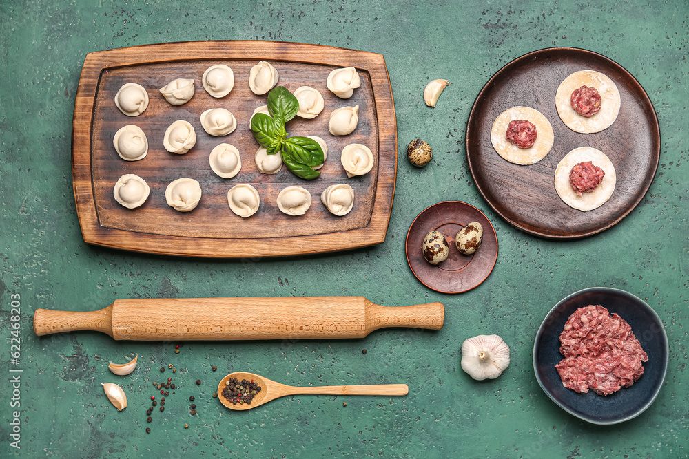 Wooden board with uncooked dumplings and ingredients on green background