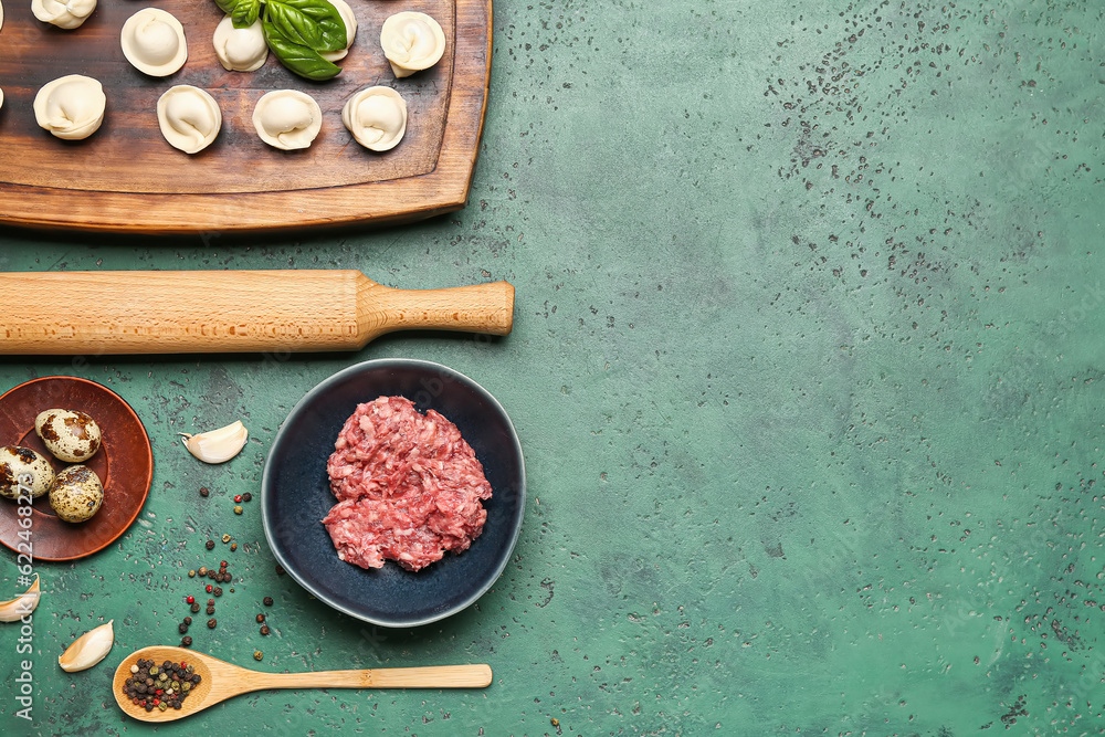 Wooden board with uncooked dumplings and ingredients on green background