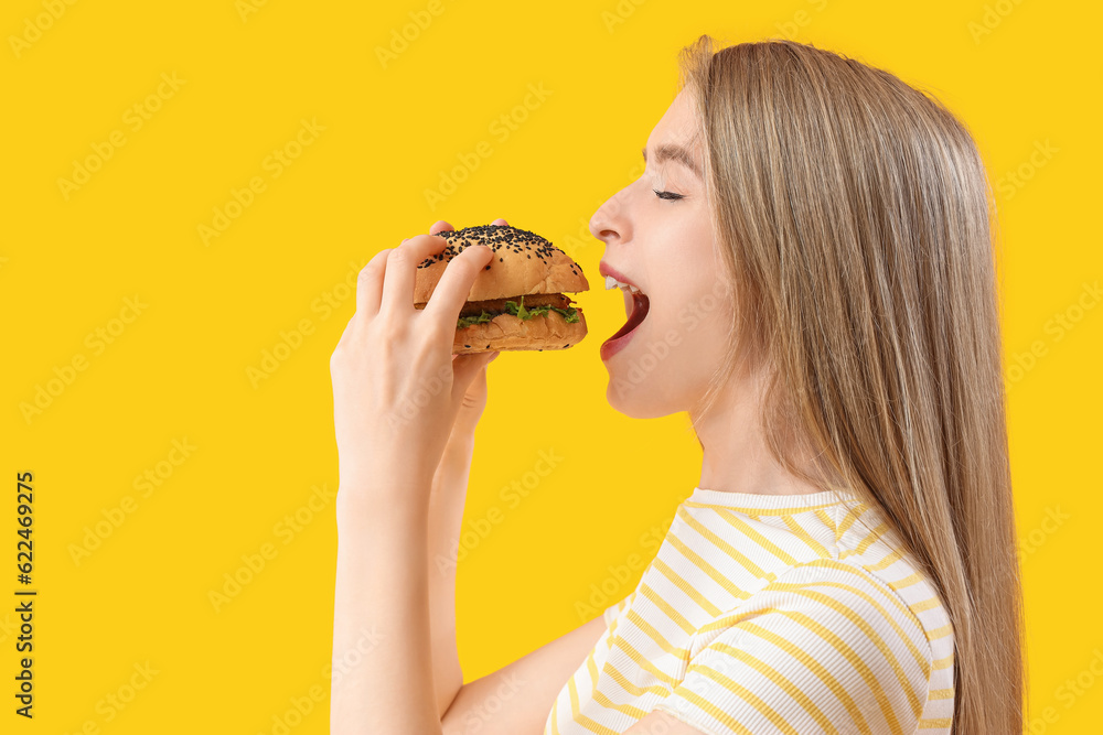 Young woman eating tasty burger on yellow background, closeup