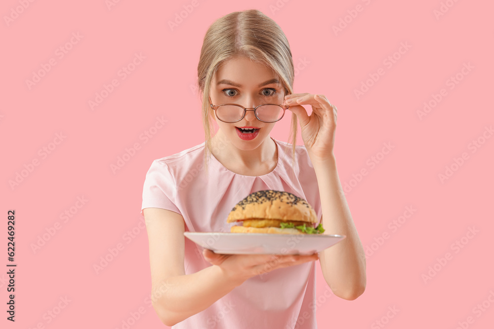 Shocked young woman with tasty burger on pink background