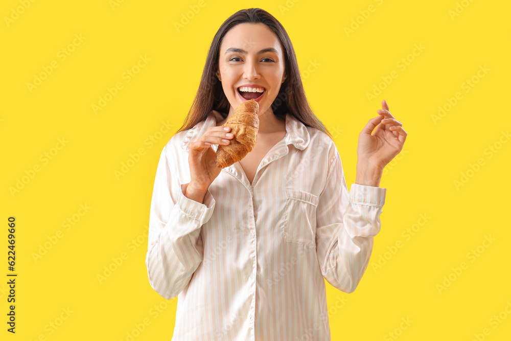 Young woman in pajamas with tasty croissant on yellow background