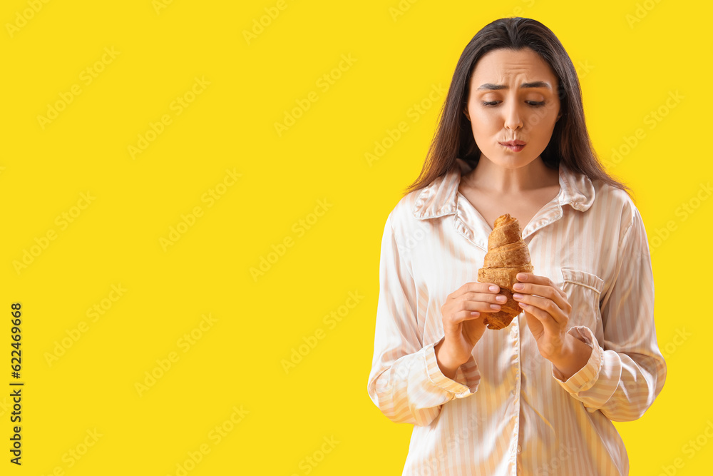 Worried young woman in pajamas with tasty croissant on yellow background