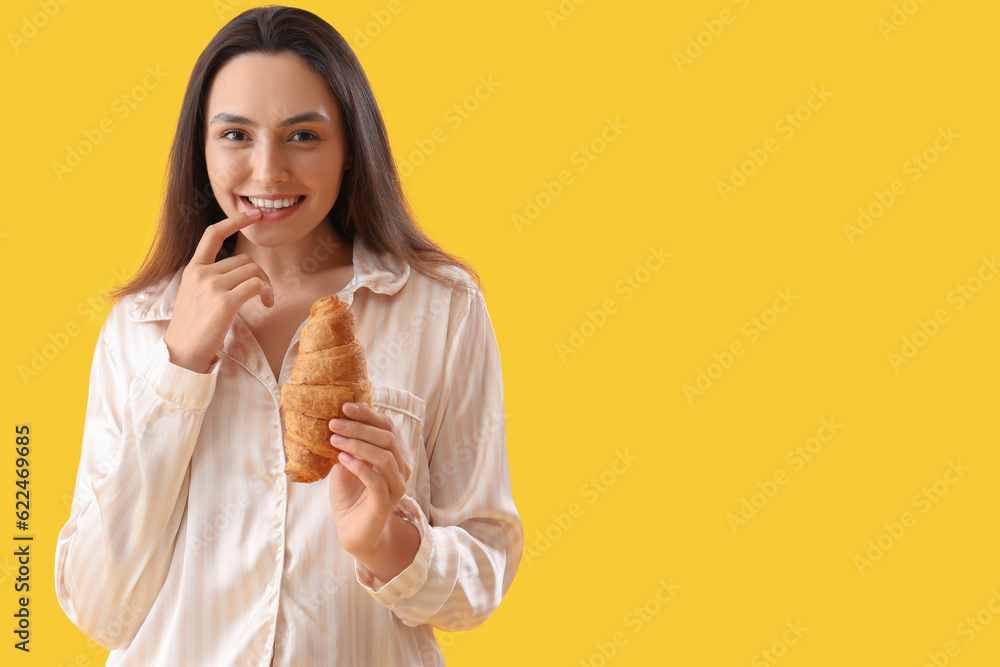 Young woman in pajamas with tasty croissant on yellow background