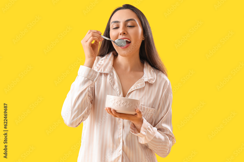 Young woman in pajamas with spoon and bowl on yellow background