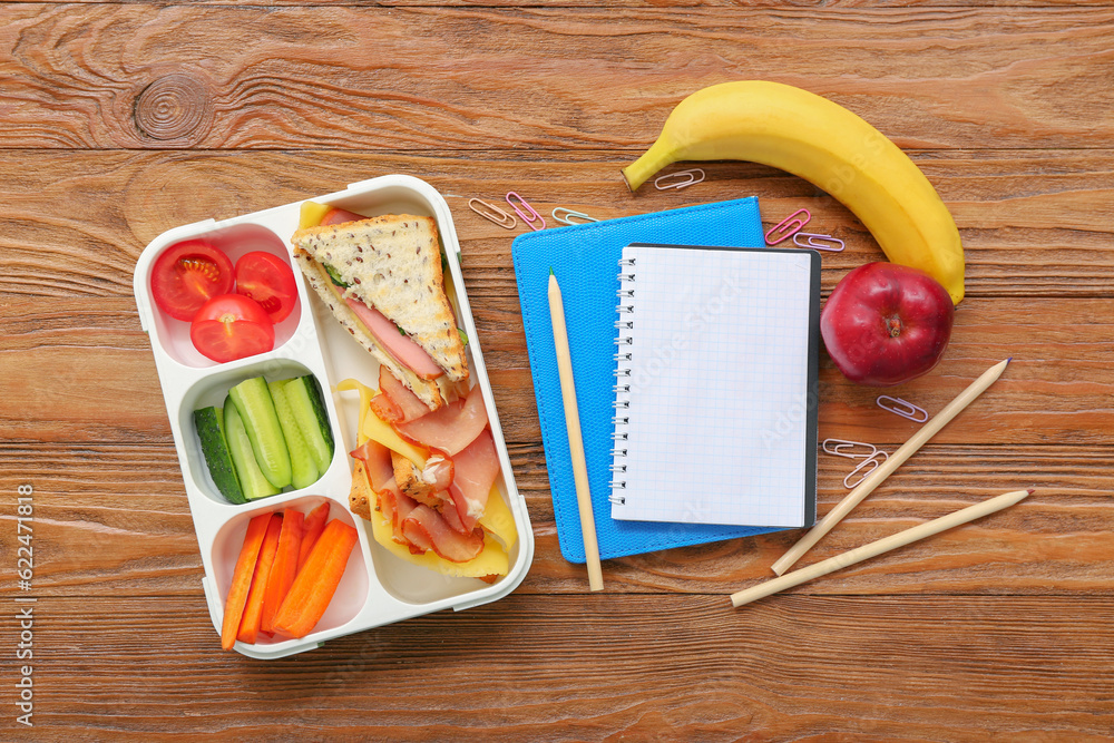 Stationery and lunch box with tasty food on brown wooden background