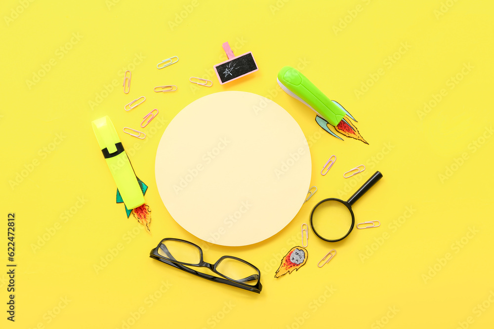 Creative composition with blank card, paper rockets and different stationery on yellow background