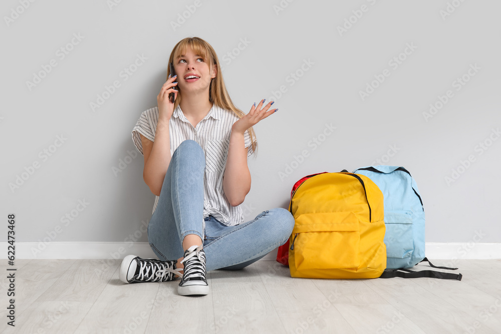 Female student with backpacks talking by mobile phone near light wall