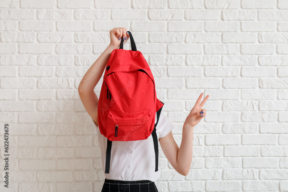 Female student with backpack showing victory gesture on white brick background