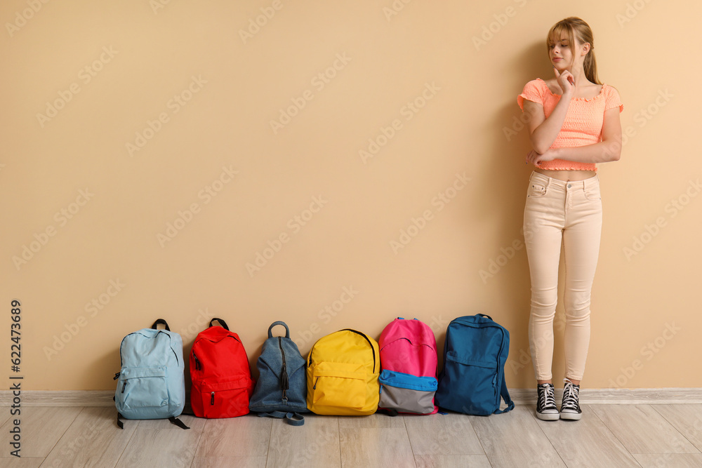 Thoughtful female student with backpacks near beige wall