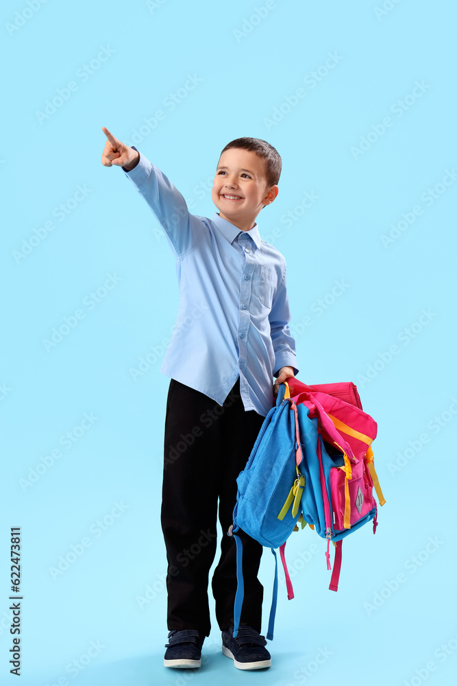 Little schoolboy with backpacks pointing at something on light blue background