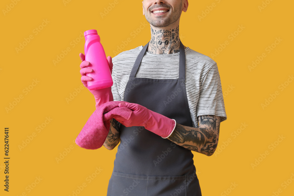 Young tattooed man with bottle of detergent and rag on orange background, closeup