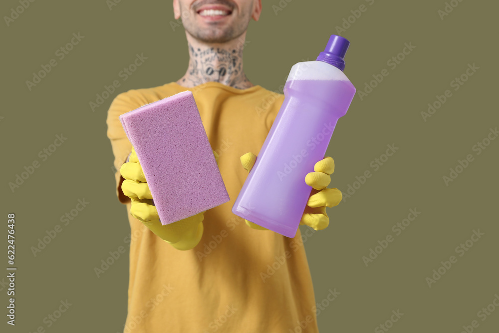 Young tattooed man with bottle of detergent and cleaning sponge on green background, closeup