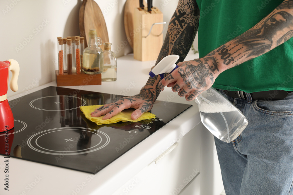 Young tattooed man cleaning electric stove in kitchen, closeup