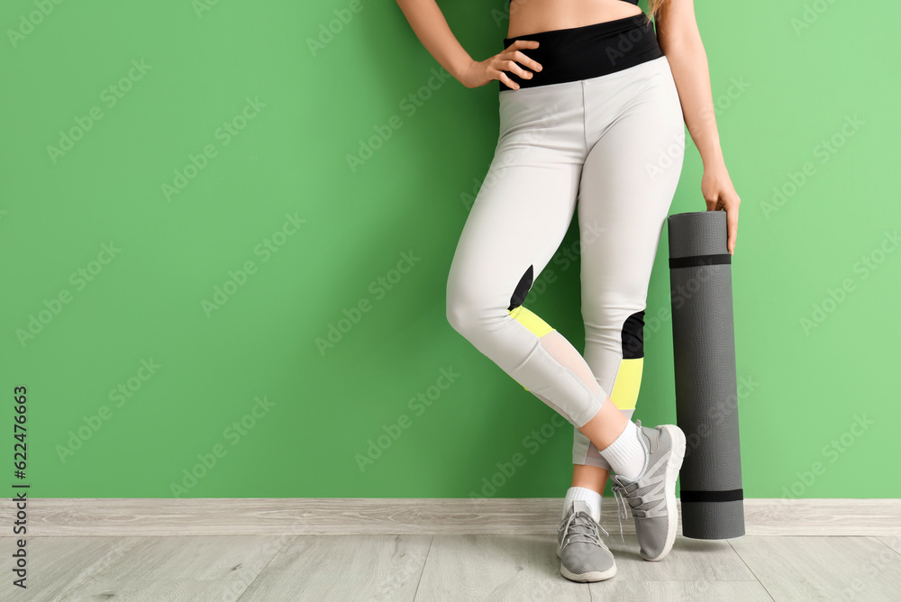 Young woman in sportswear and with yoga mat near green wall