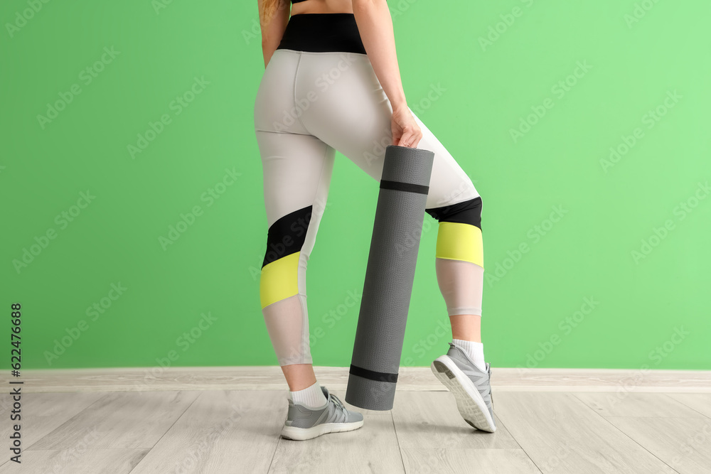 Young woman in sportswear and with yoga mat near green wall