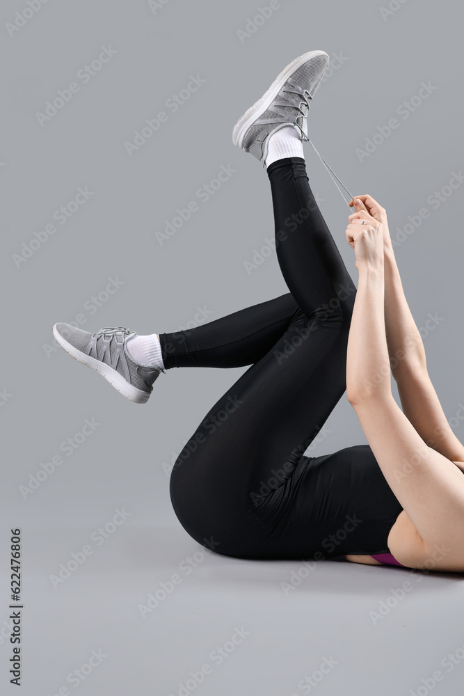 Young woman in sportswear tying shoelaces on grey background