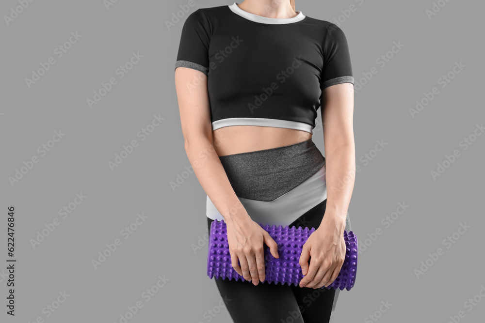 Young woman in sportswear and with foam roller on grey background