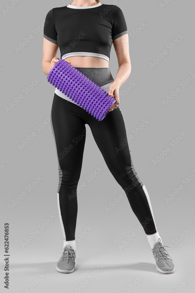 Young woman in sportswear and with foam roller on grey background