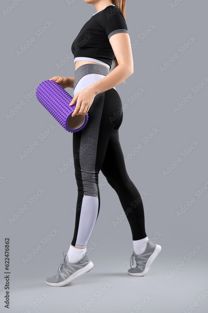 Young woman in sportswear and with foam roller on grey background