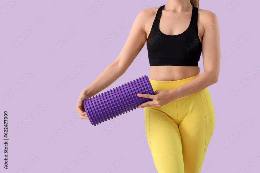 Young woman in sportswear and with foam roller on lilac background